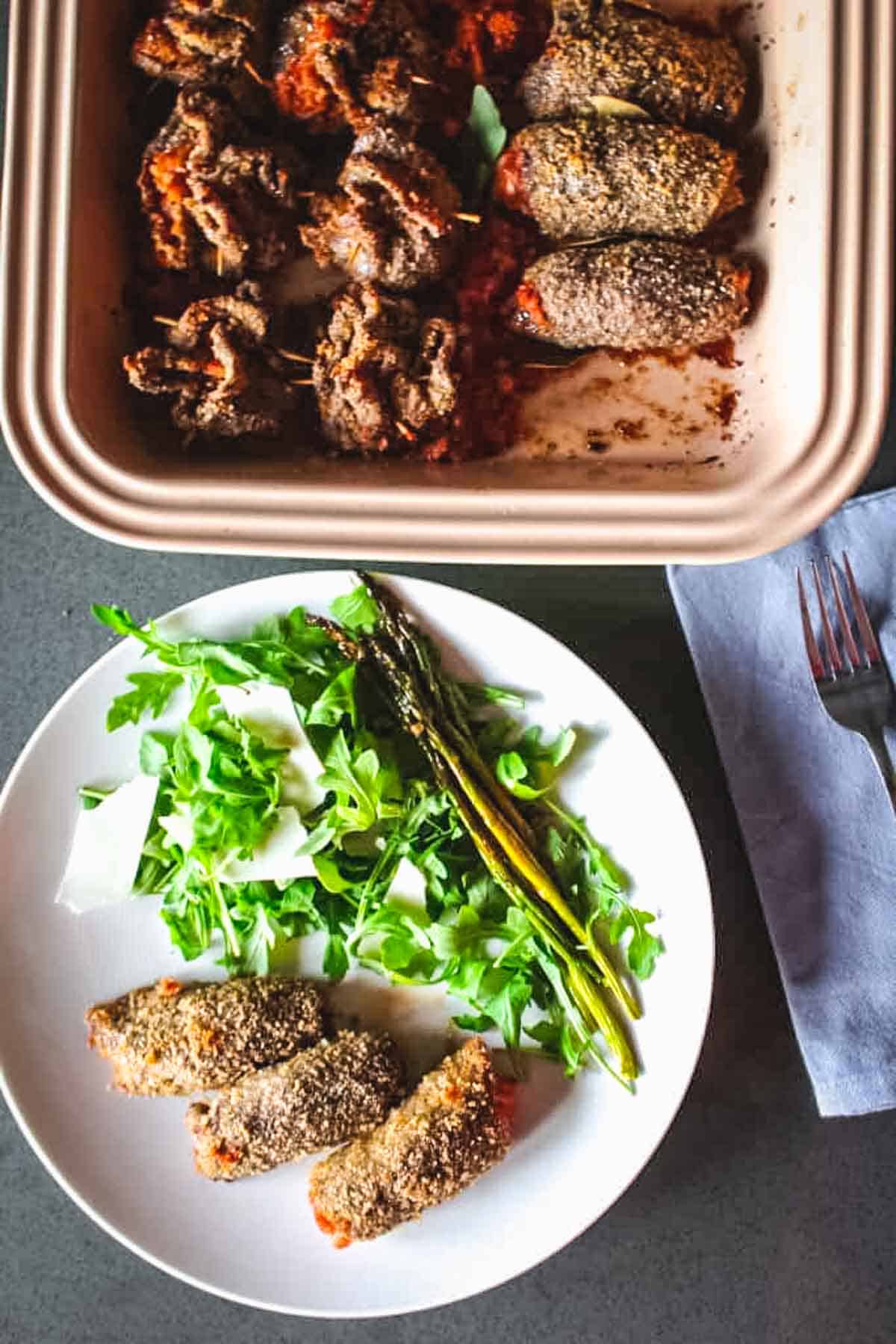 A white plate with Sicilian spiedini, arugula salad and asparagus.