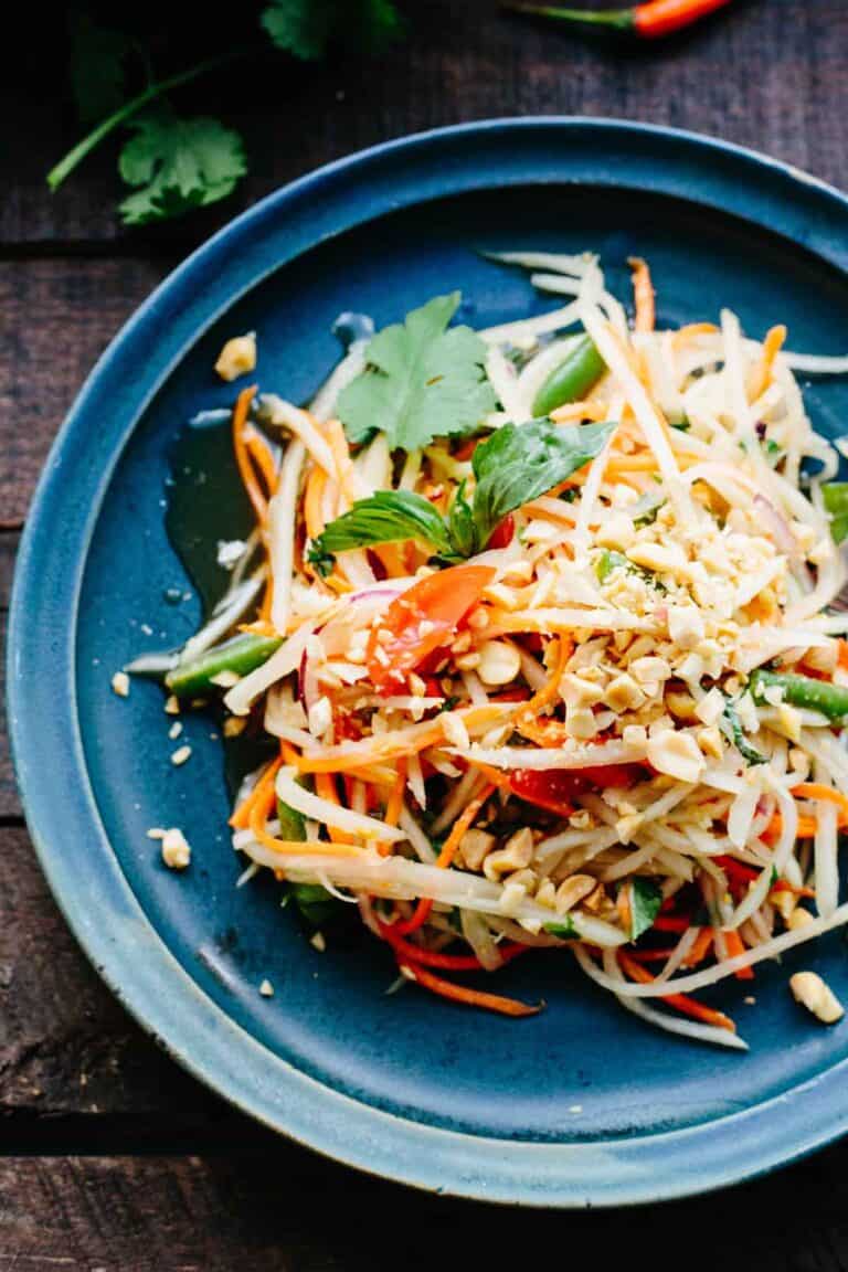 Thai papaya salad with different colored vegetables and herbs on a blue plate.