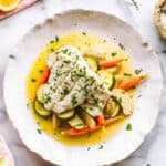 A white plate with white fish over vegetables in a butter sauce.