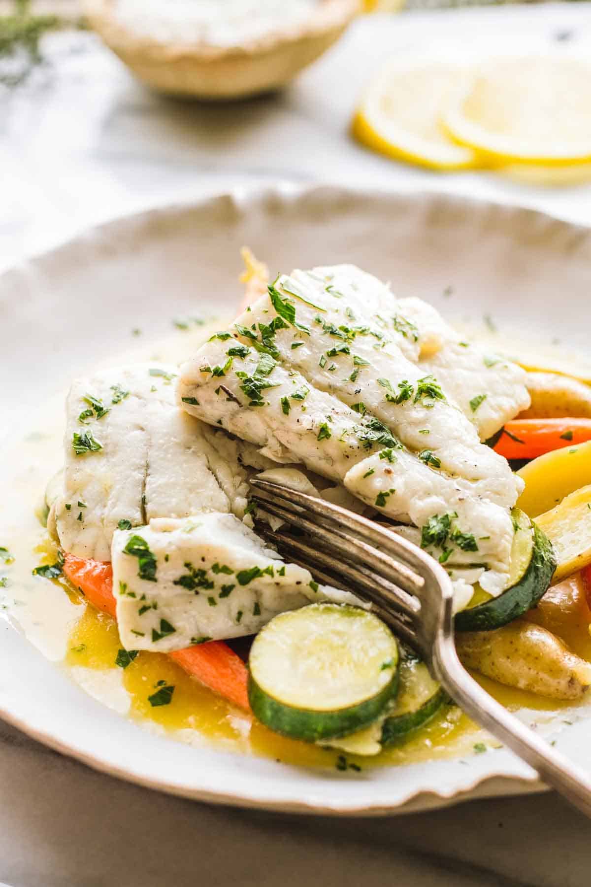 A fork breaking through a piece of tender flaky white fish with parsley on top and vegetables beneath.