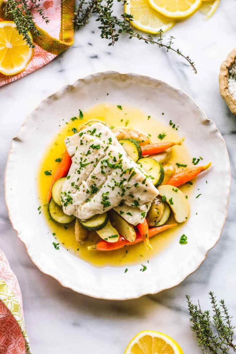 A white plate with white fish over vegetables in a butter sauce.