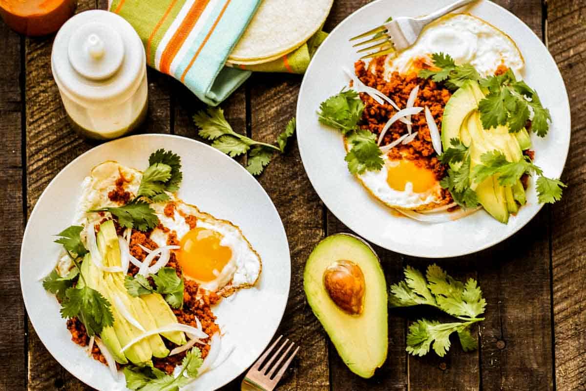 Two plates of chorizo and eggs with avocado, hot sauce and cilantro.
