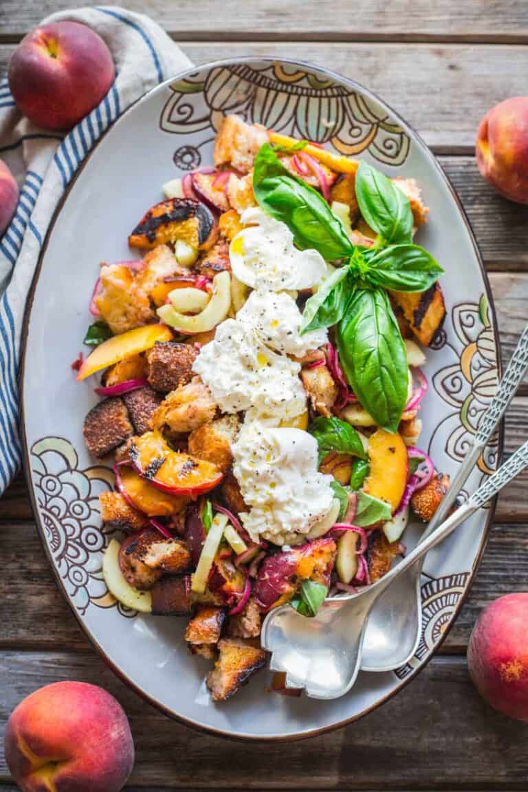 A serving dish of grilled peach and bread salad with burrata and serving spoons.
