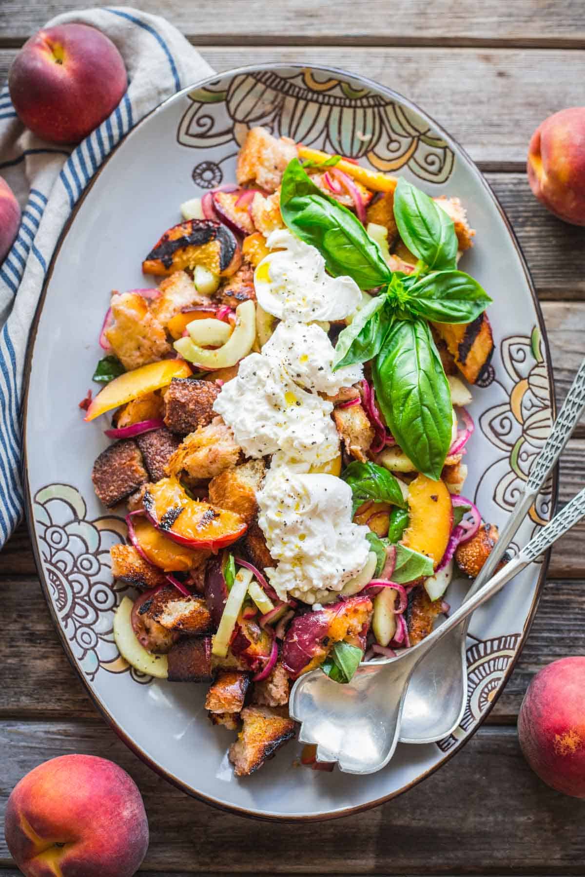 A serving dish of grilled peach and bread salad with burrata and serving spoons.