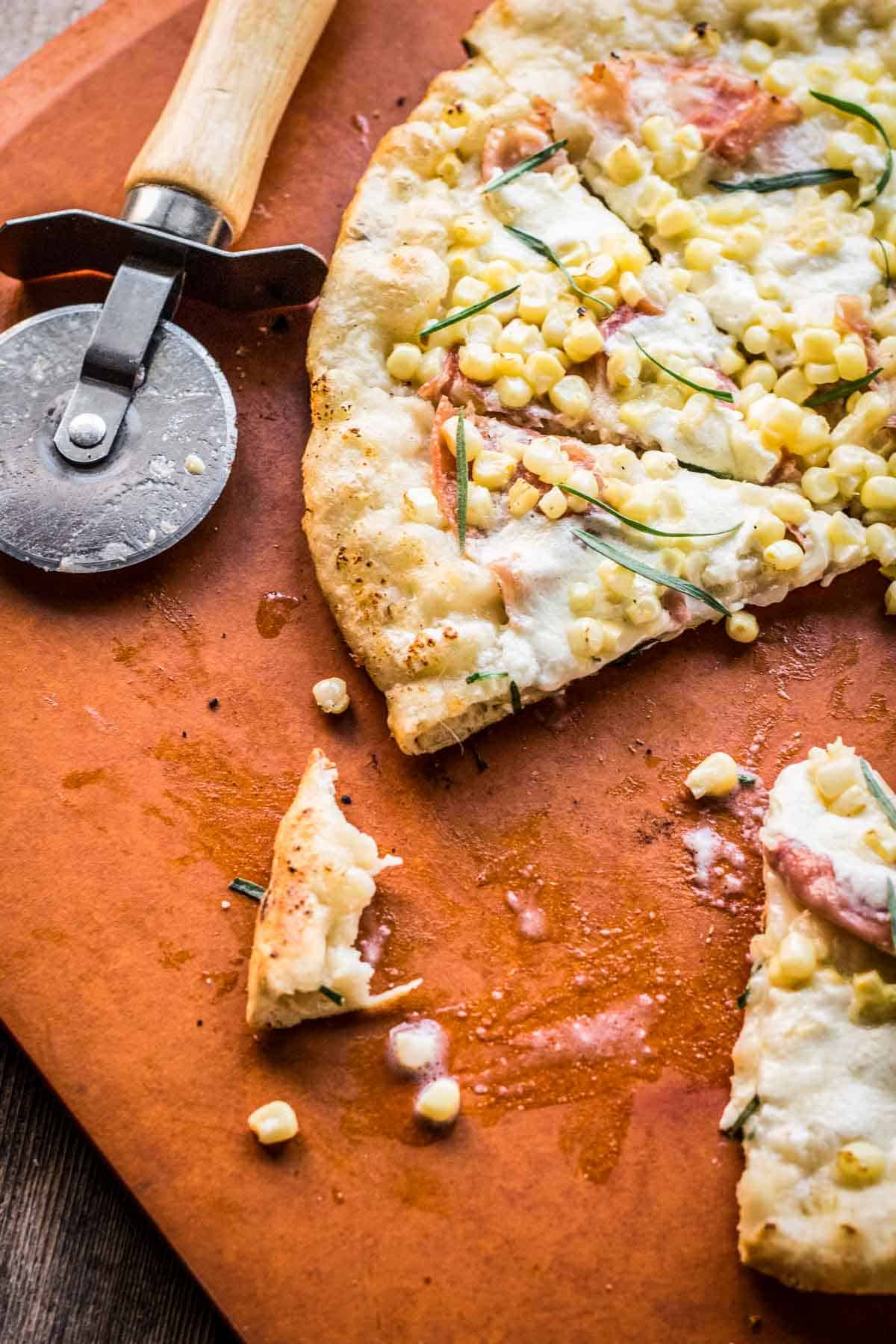 A grilled corn pizza cut into wedges with a pizza cutter.