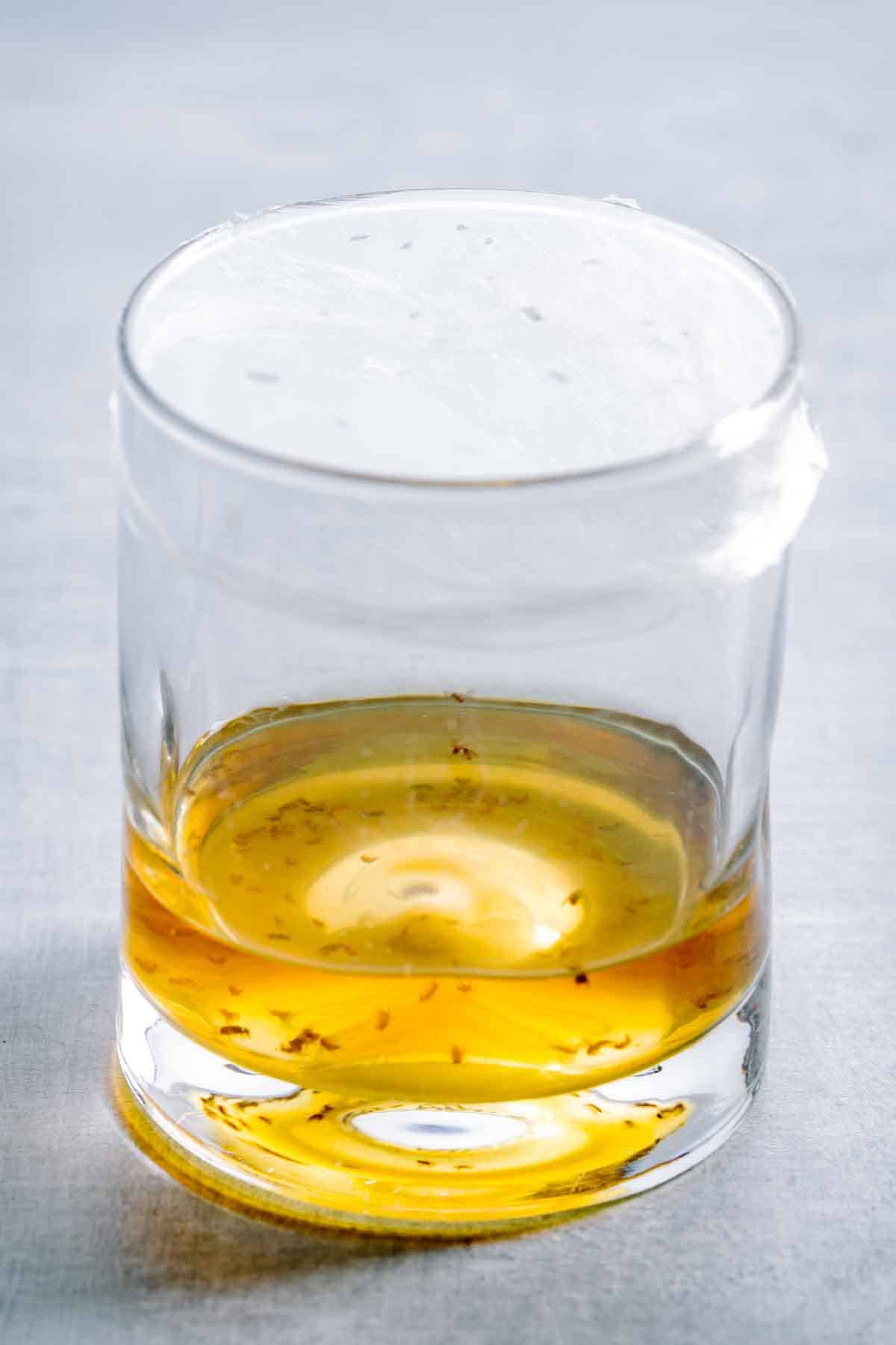 A glass filled with amber liquid and small bugs with plastic wrap on top.