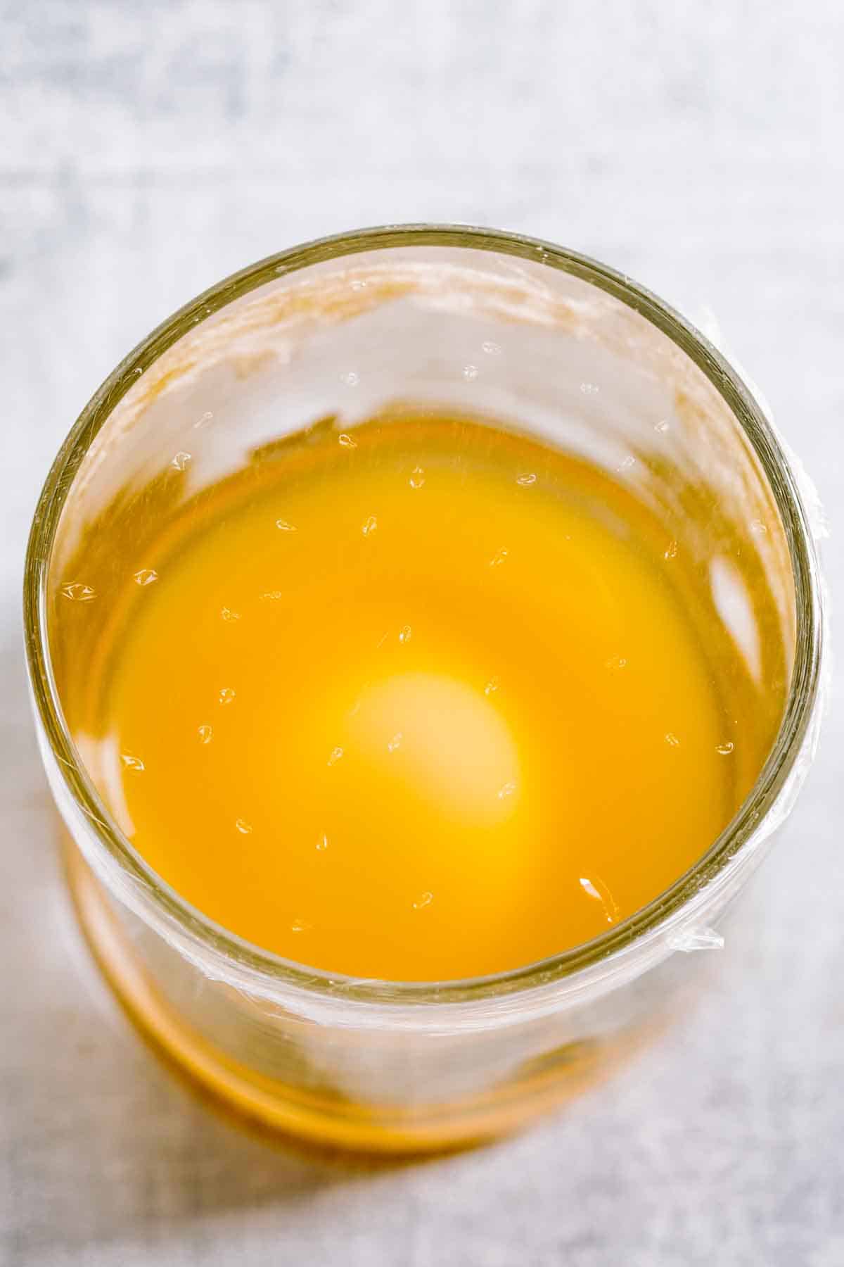 A glass with apple cider vinegar and plastic wrap with holes poked in it on top.
