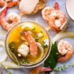 A jar of pickled shrimp surrounded by pieces of shrimp and herbs.