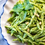 A plate of pesto pasta garnished with pine nuts and a sprig of basil.