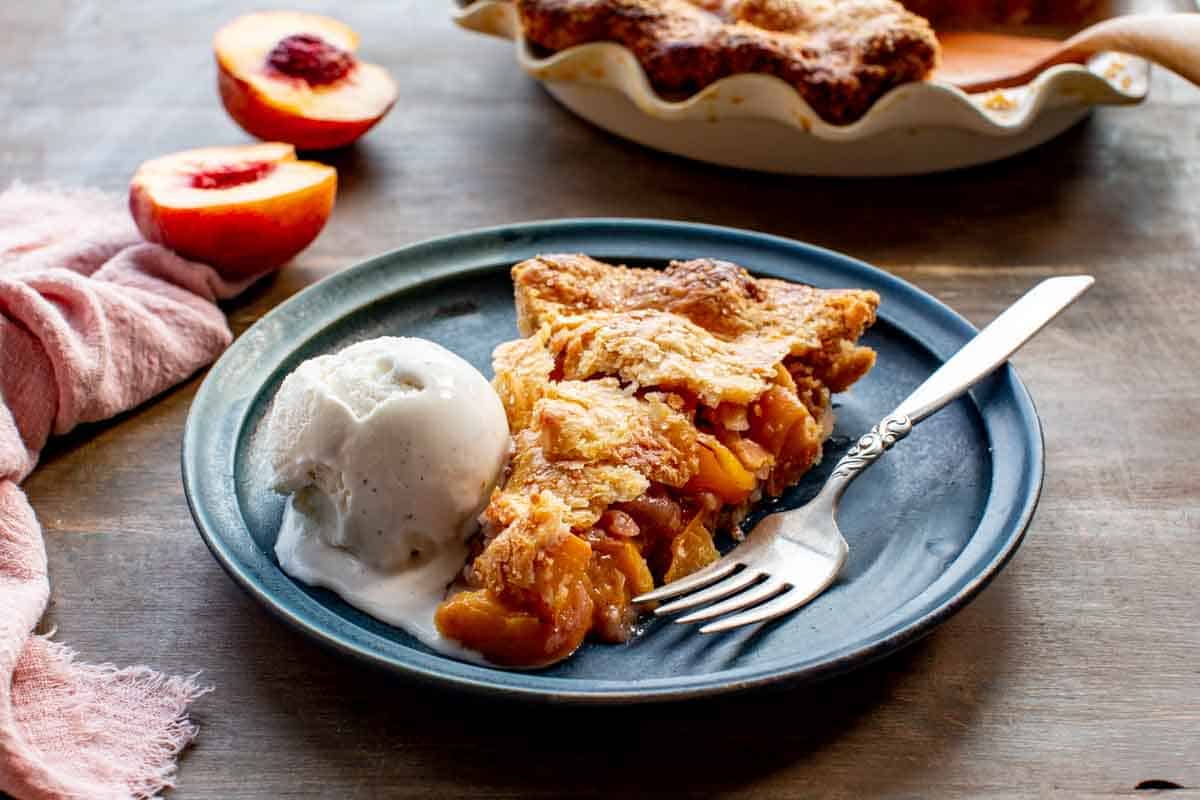 A blue plate with a slice of peach pie and a scoop of vanilla ice cream with a fork.