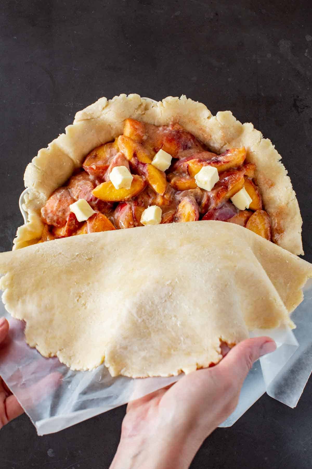 Placing a top crust over top of peaches and butter in a pie shell.