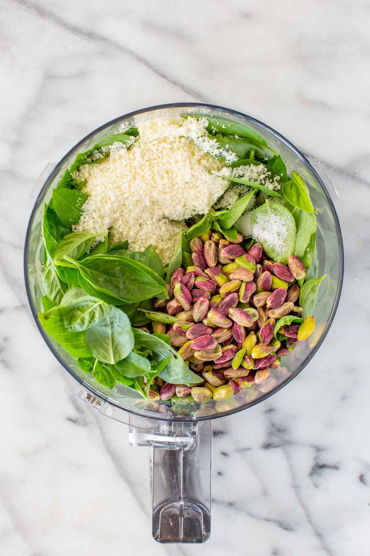 A food processor with fresh basil, grated cheese and shelled pistachios.