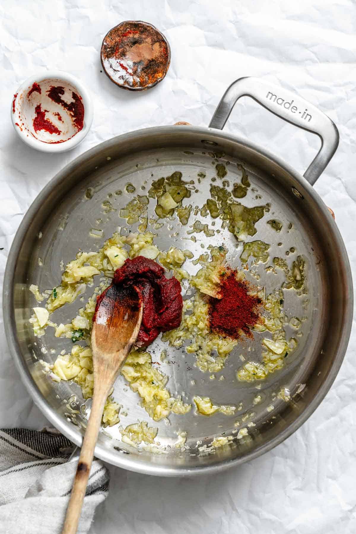 A wooden spoon stirring tomato past and smoked paprika into sautéed garlic and shallots.