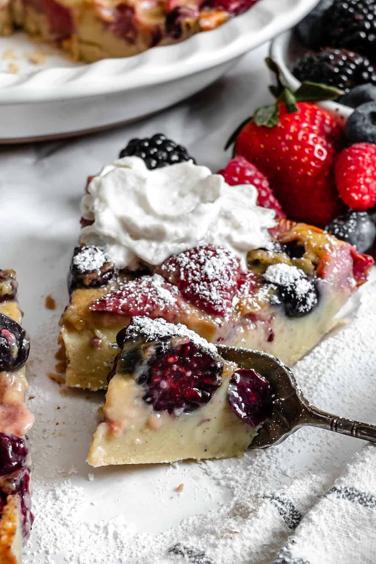 View of berry clafoutis with a slice removed, served with fresh berries and whipped cream.