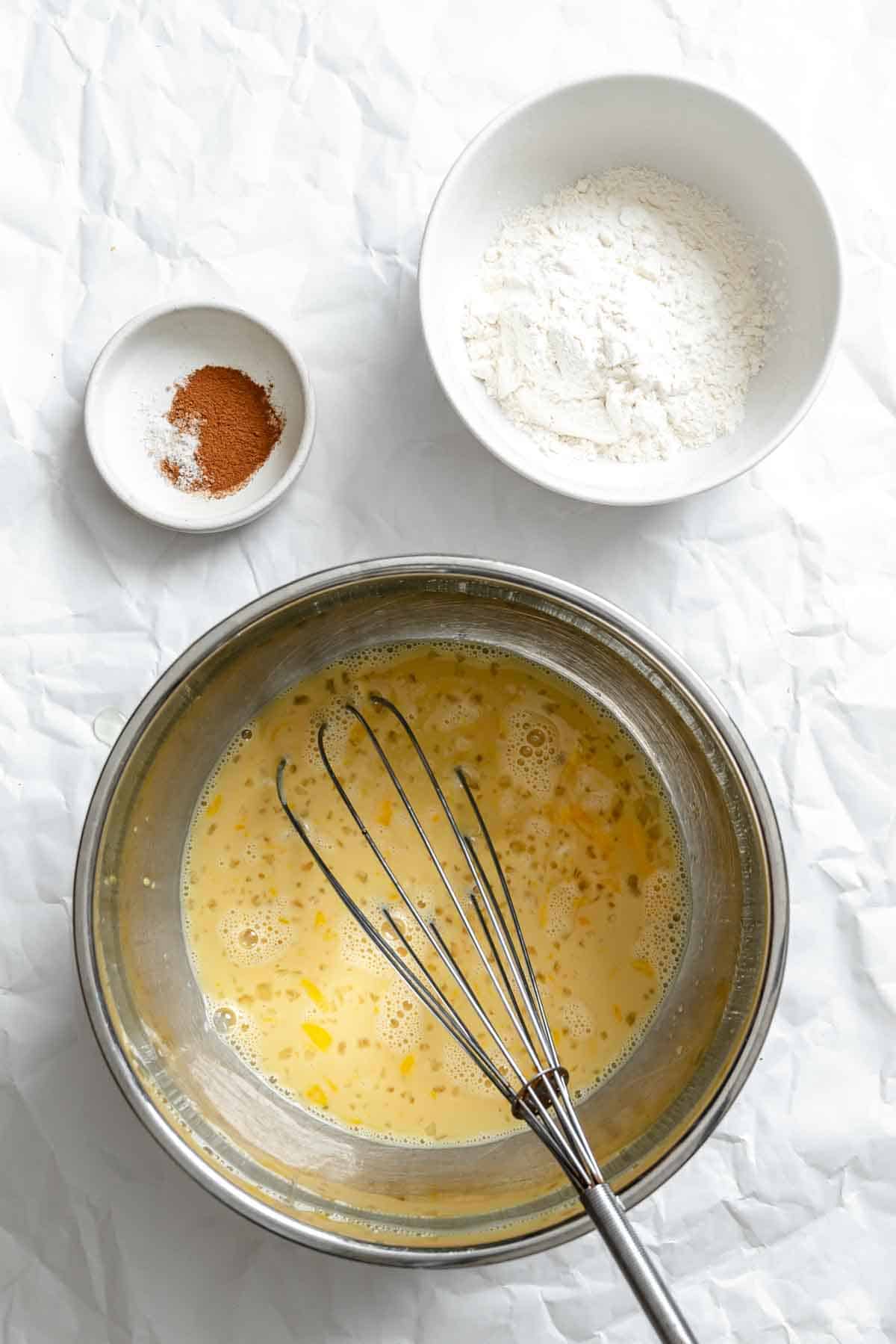 Bowl of flour next to a small dish of cinnamon and a metal bowl containing whisked egg mixture.