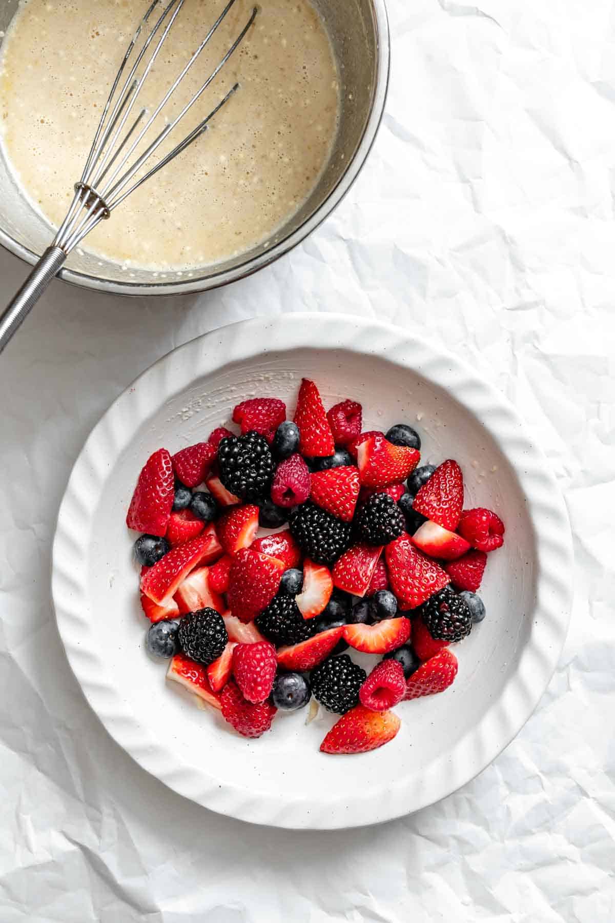 Ceramic baking dish with berries next to a metal bowl containing whisked egg mixture.