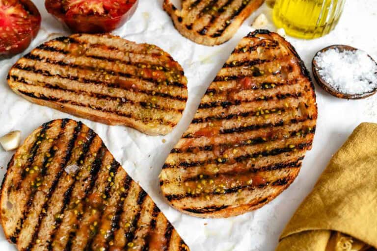 More grilled bread slices on parchment paper with tomatoes, olive oil, and a jar of olive oil in the corner.