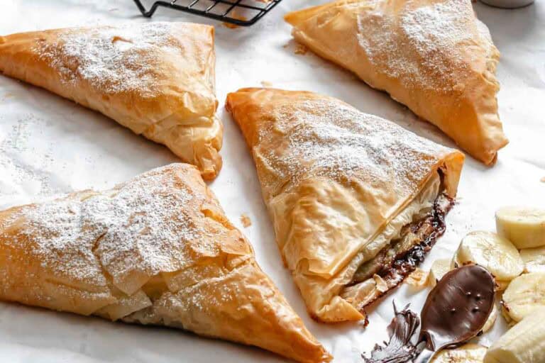 Stacked broken phyllo triangles revealing Nutella filling, dusted with sugar and surrounded by bananas.