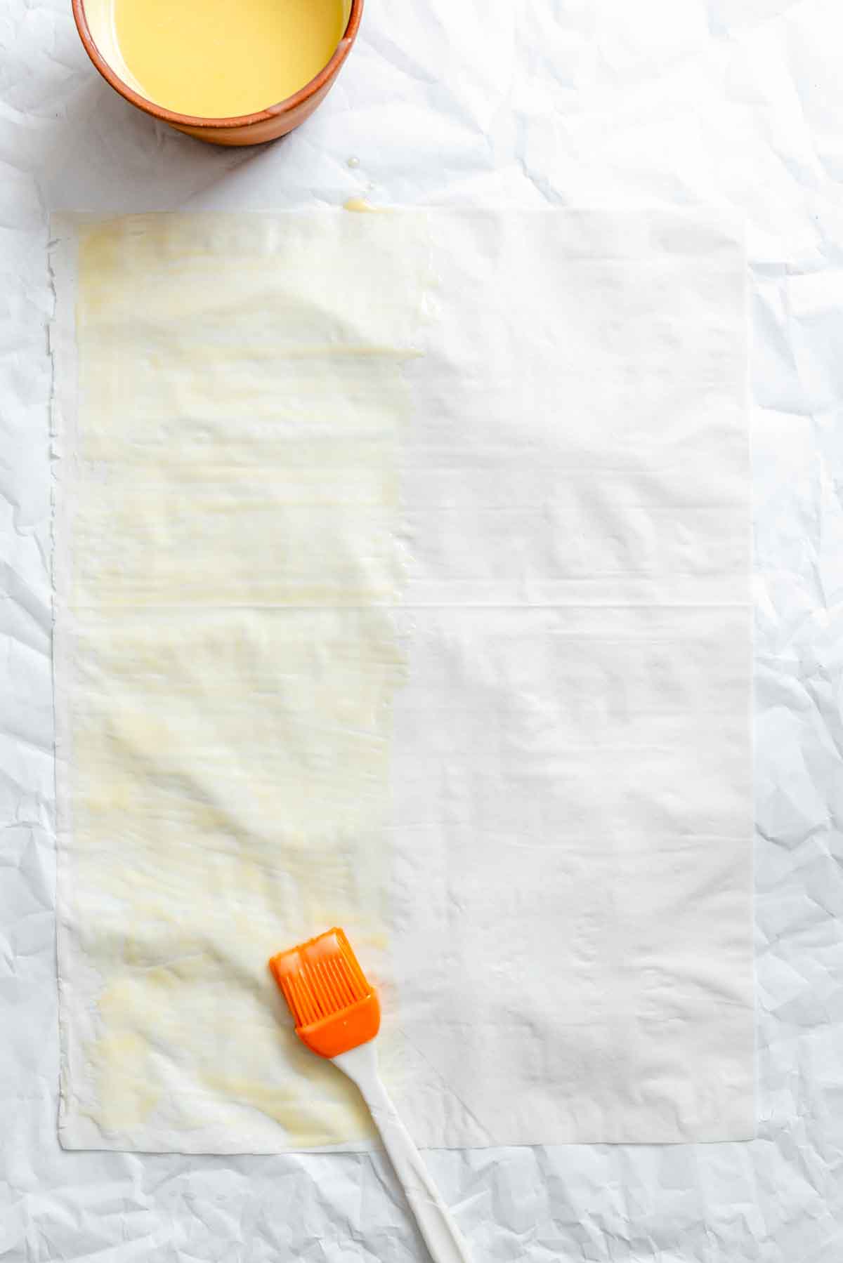 Phyllo dough sheet on parchment with a bowl of butter and orange pastry brush applying butter to one edge.