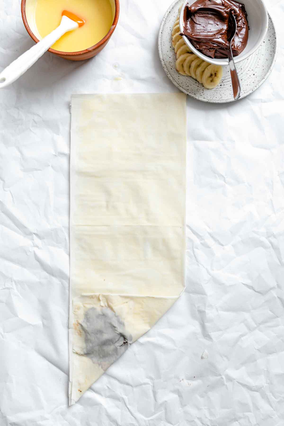 Folded phyllo dough with bowls of butter and Nutella, and sliced bananas on a plate.