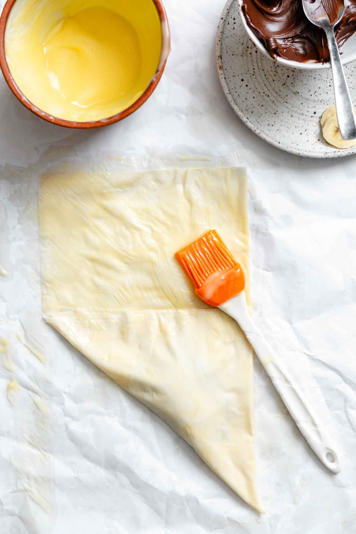 Triangular phyllo dough with pastry brush applying butter, bowls of ingredients visible.
