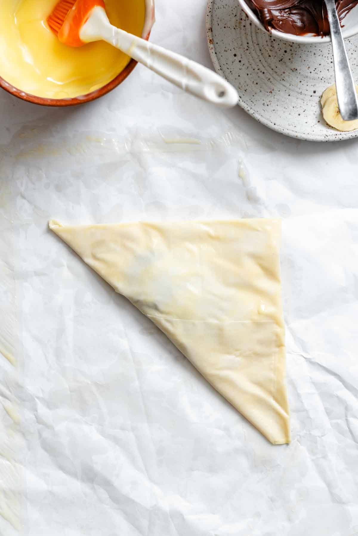 Folded phyllo triangle with pastry brush, beside bowls of butter and Nutella with bananas.