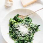 A white bowl of sautéed spinach topped with freshly grated Parmigiano-Reggiano cheese and a silver spoon, with a head of garlic and slice of cheese visible in the background.