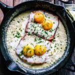 An overhead view of a cast iron skillet containing two slices of ham topped with four eggs (two on each slice), all baking in a creamy sauce and sprinkled generously with fresh chives. Cracked eggshells and fresh chives are visible in the background, emphasizing the dish's homemade quality.