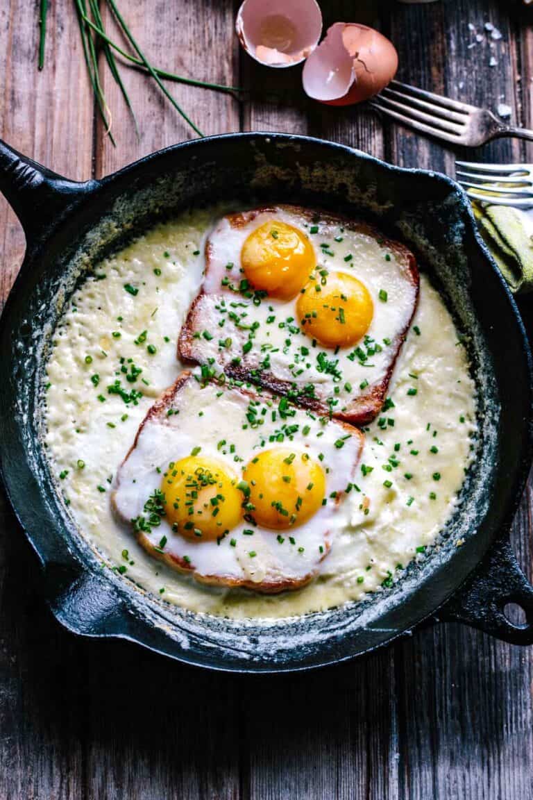 An overhead view of a cast iron skillet containing two slices of ham topped with four eggs (two on each slice), all baking in a creamy sauce and sprinkled generously with fresh chives. Cracked eggshells and fresh chives are visible in the background, emphasizing the dish's homemade quality.