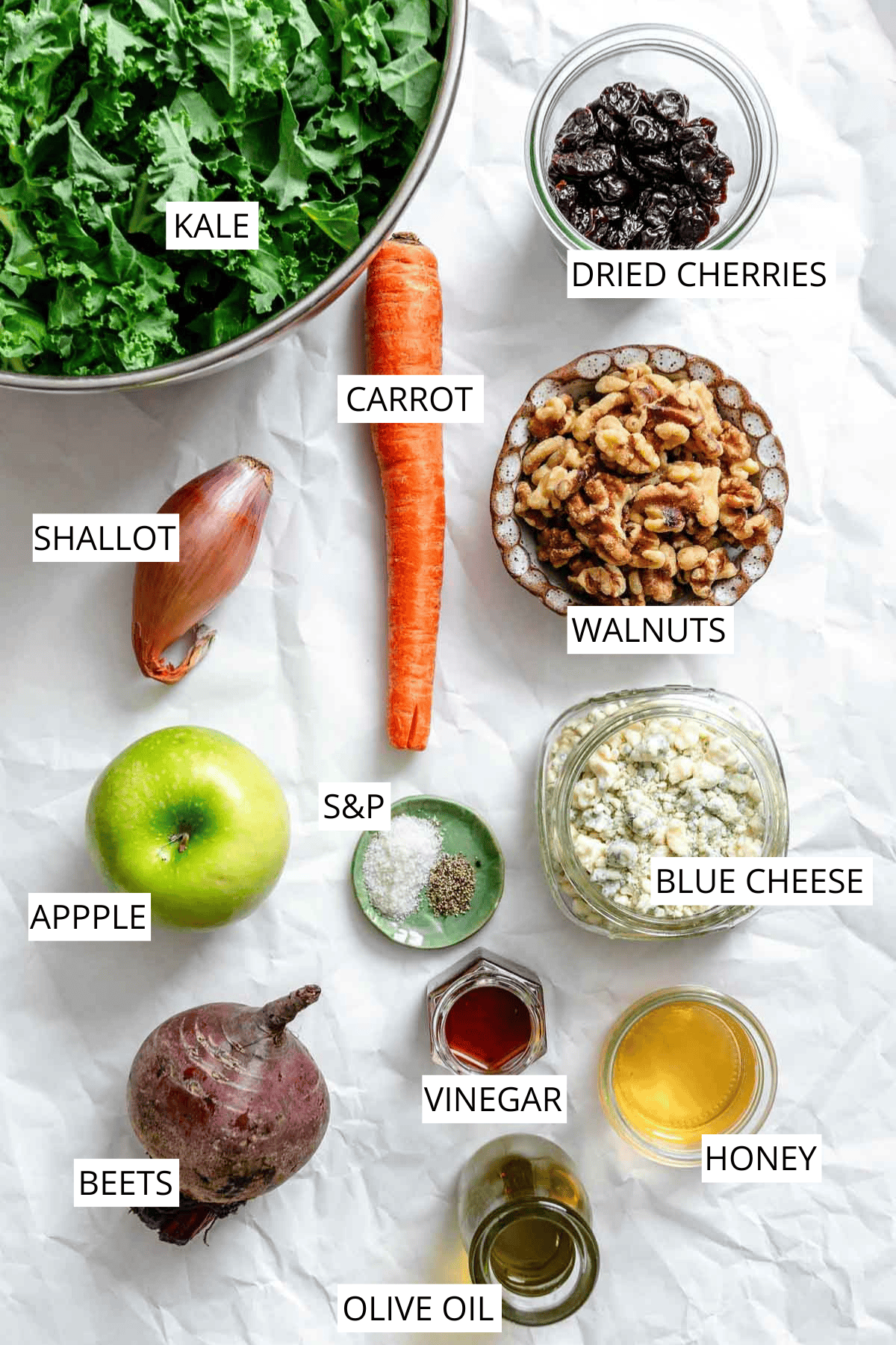 An ingredient layout for the fall harvest salad featuring a bowl of fresh kale, carrot, apple, beet, shallot, containers of dried cranberries, walnuts, blue cheese, and small jars of various dressings and oils arranged on white parchment paper.