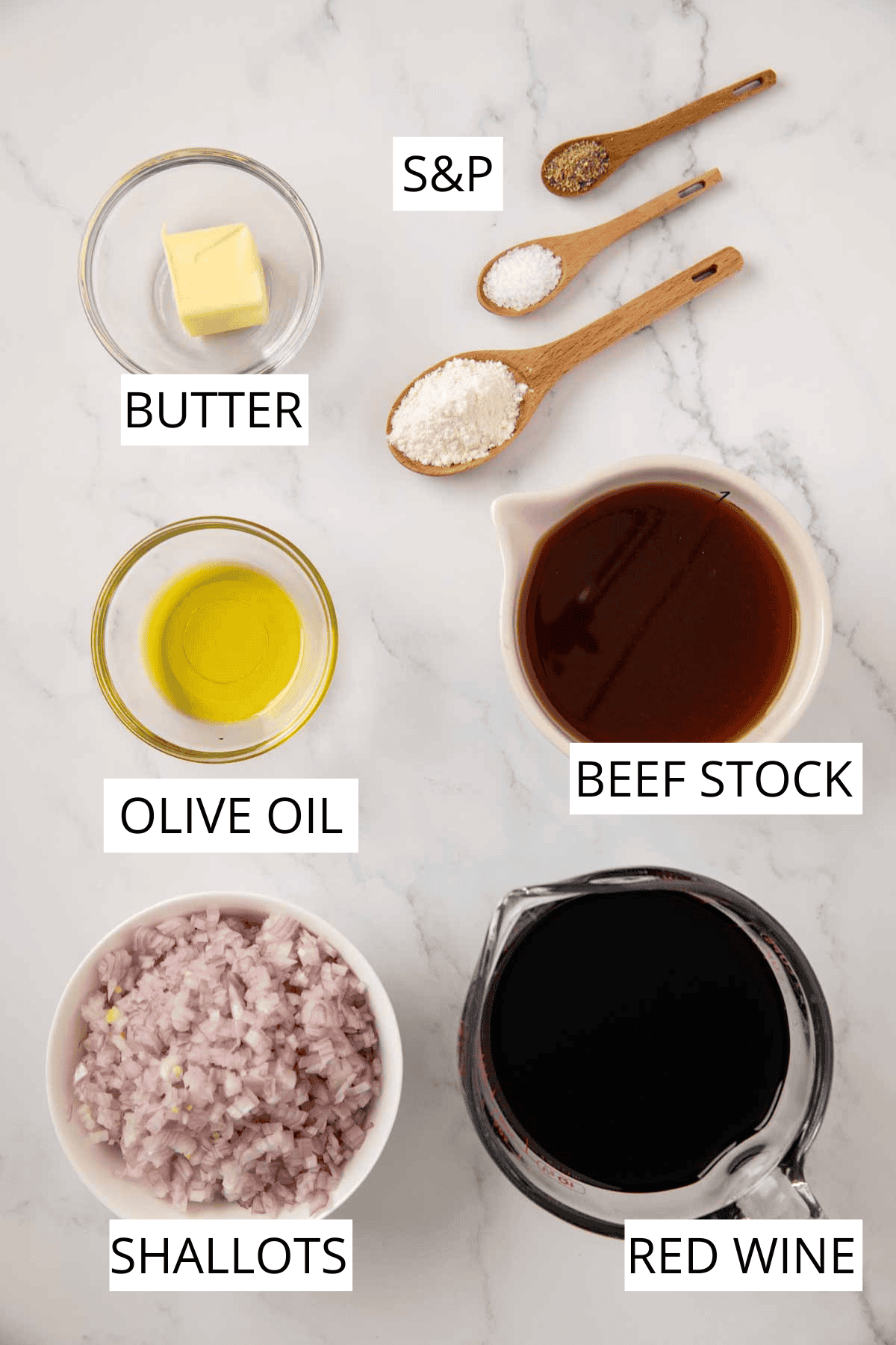 A flat-lay of ingredients for bordelaise sauce on a marble countertop, including butter, olive oil, wooden spoons with salt and flour, beef stock in a white pitcher, minced shallots in a small bowl, and red wine in a glass measuring cup.