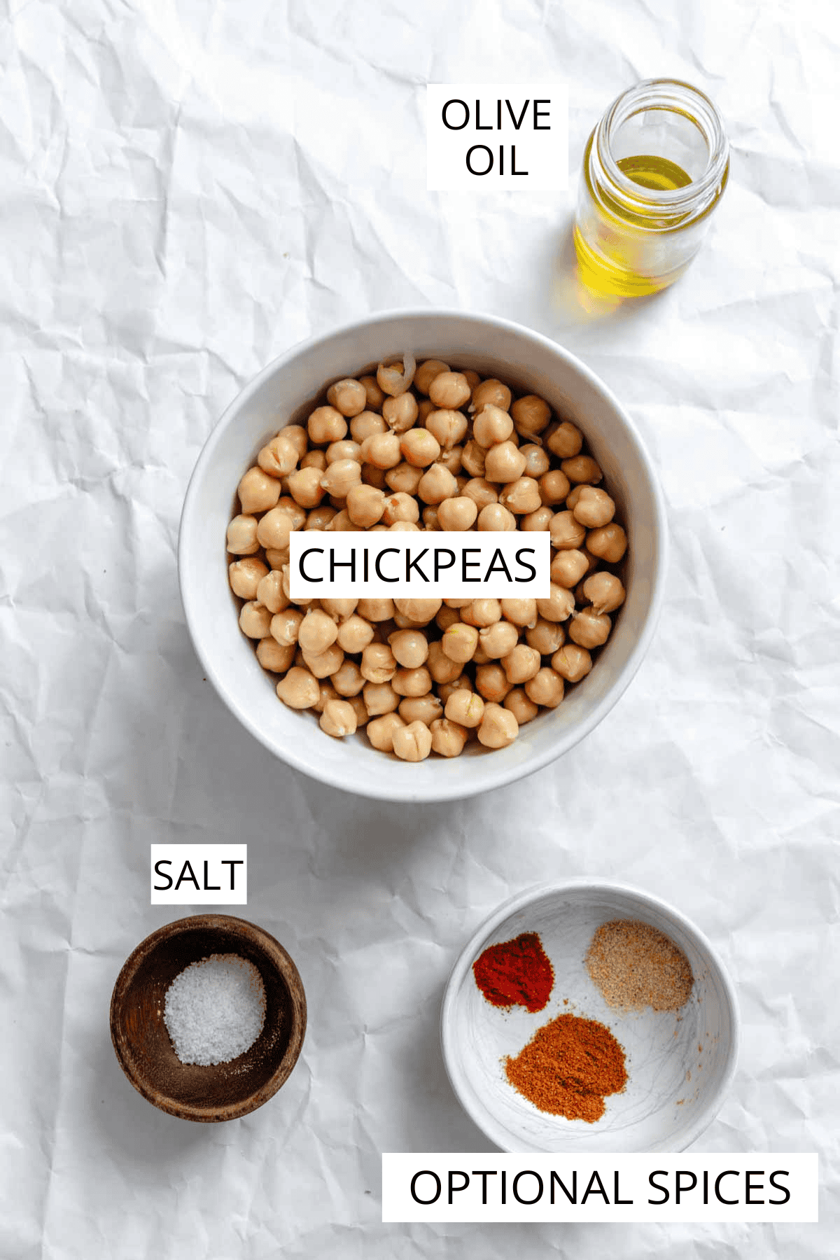 Overhead view of raw chickpeas in a white bowl with small bowls of spices and a jar of olive oil against white parchment background.