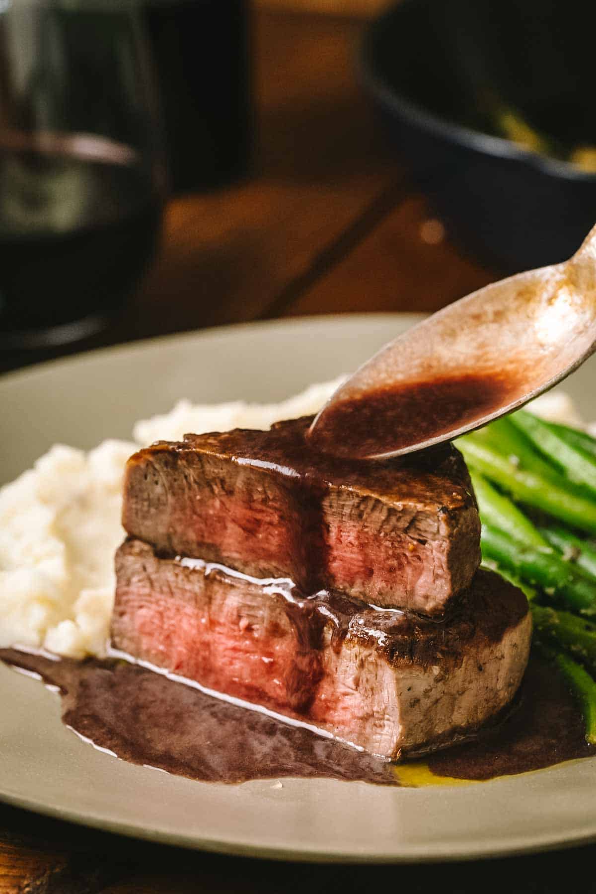 Another angle of bordelaise sauce being drizzled over sliced steak medallions arranged on a plate with mashed potatoes and green beans, highlighting the glossy, dark brown color of the finished sauce.