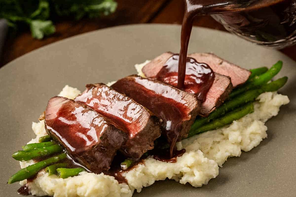 Sliced steak on a bed of mashed potatoes with bordelaise sauce being poured from a small glass pitcher, showing the sauce flowing smoothly over the meat on a white plate.
