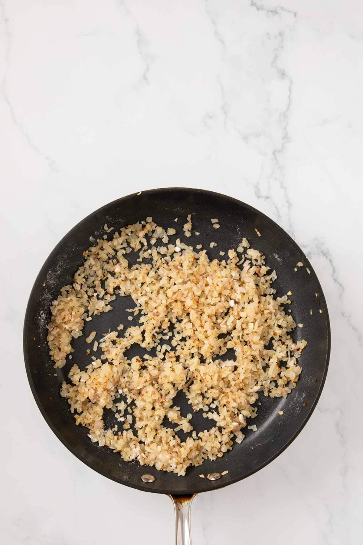 A black cast iron pan on a white marble countertop containing sautéed minced onions or shallots, which appear golden brown and evenly distributed across the cooking surface.