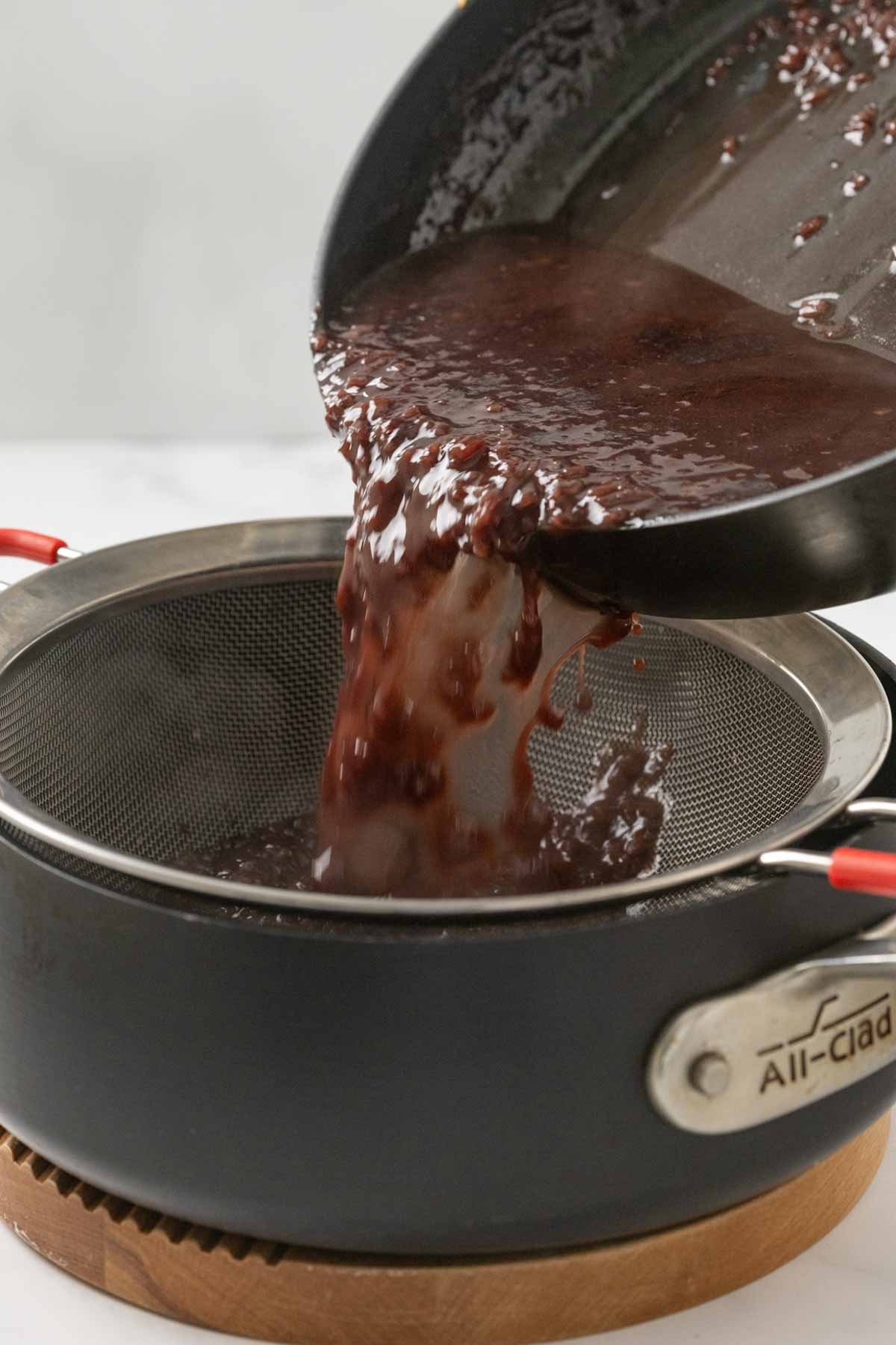 A close-up action shot of dark red-brown sauce being poured from a black skillet through a fine mesh strainer into what appears to be a saucepan. The liquid is being strained to remove solid ingredients.