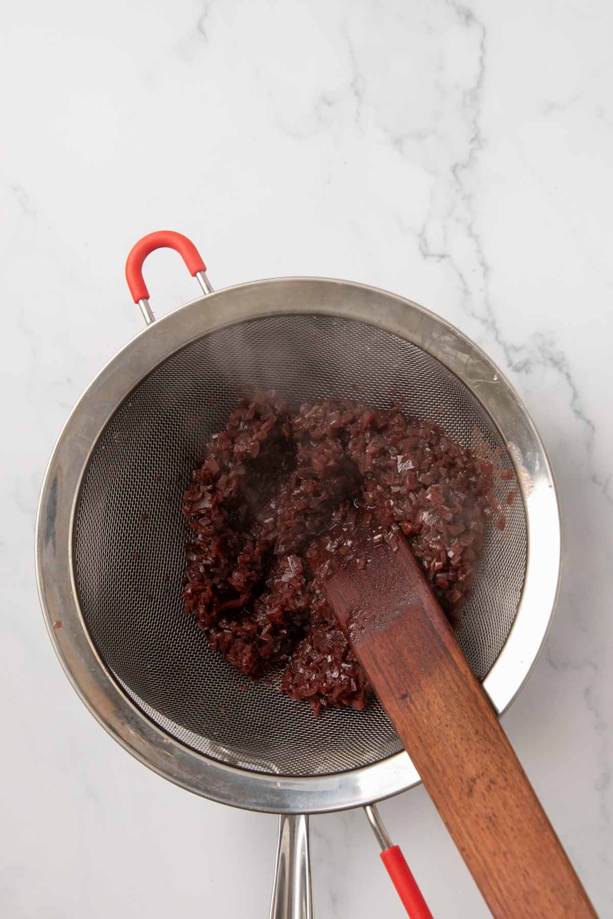 A top-down view of a fine mesh strainer containing strained solids from the sauce, with a wooden spoon pressing down to extract all liquid. The strainer rests over what appears to be a saucepan on a white marble surface.