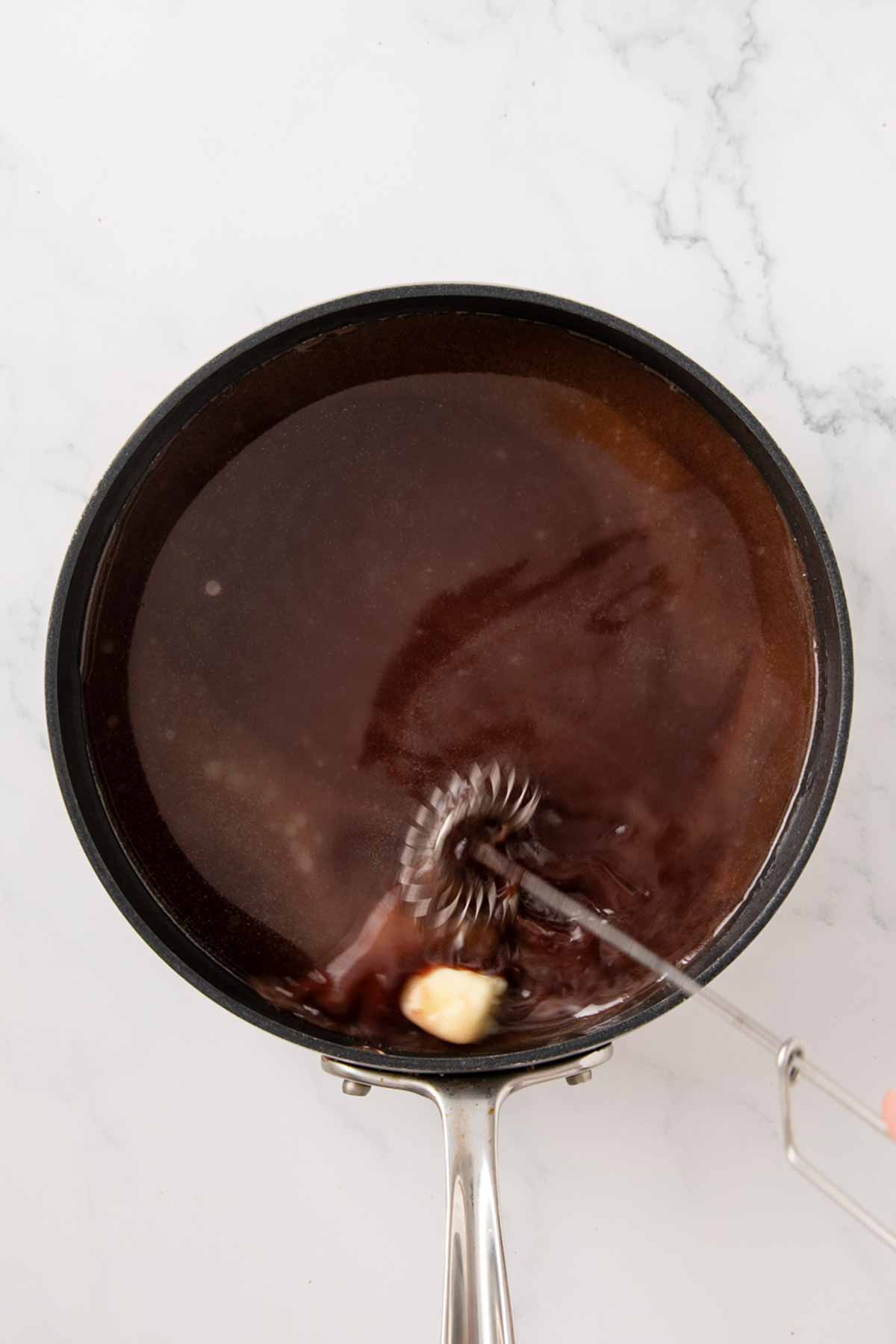 An overhead view of a pan containing dark bordelaise sauce with a metal whisk visible, showing the sauce being emulsified with what appears to be butter, creating a glossy, rich texture.