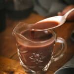 A glass measuring cup filled with rich brown bordelaise sauce, with a spoon drizzling some of the sauce back into the cup. The sauce has a thick, velvety consistency characteristic of a well-reduced sauce.