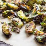 A closer view of roasted Brussels sprouts on parchment paper, showing their golden-brown exterior. The vegetable pieces are tossed with what appears to be finely grated Pecorino cheese and fresh herbs, giving them a textured coating.