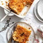 A serving of parsnip and celery root gratin on a white plate with a serving in the baking dish, showing the creamy, layered texture inside and the crispy top, accompanied by vintage utensils and a sprig of thyme.
