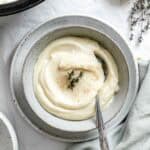 Celery root and parsnip puree served in a speckled ceramic bowl, garnished with fresh thyme and a light dusting of spice. A silver spoon rests in the creamy mixture.