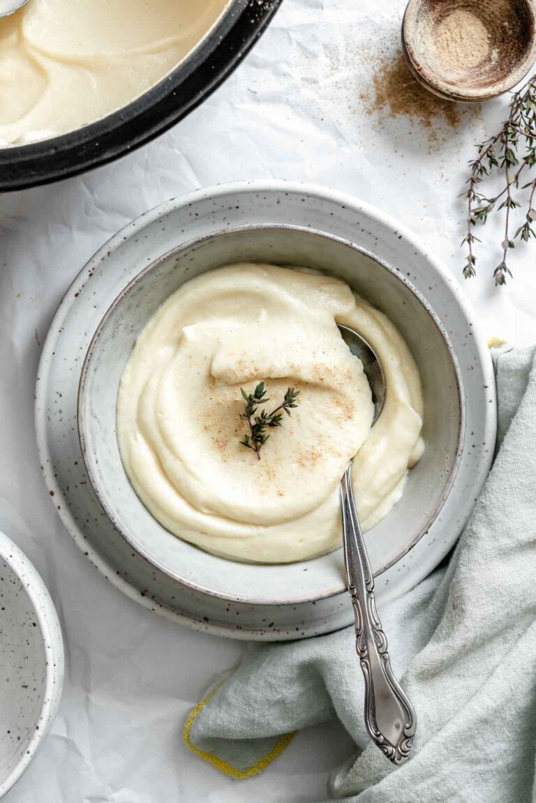 Celery root and parsnip puree served in a speckled ceramic bowl, garnished with fresh thyme and a light dusting of spice. A silver spoon rests in the creamy mixture.