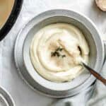 Overhead view of celery root and parsnip puree in a ceramic bowl with decorative silver spoon. The creamy white puree is garnished with fresh thyme sprigs and a light dusting of spice.