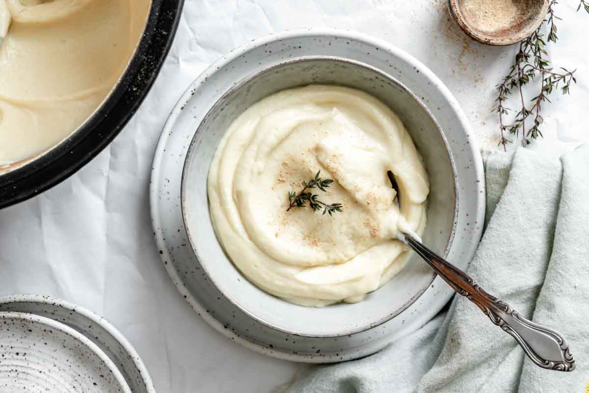 Overhead view of celery root and parsnip puree in a ceramic bowl with decorative silver spoon. The creamy white puree is garnished with fresh thyme sprigs and a light dusting of spice.