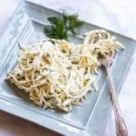 A light blue square plate containing julienned celery root remoulade with finely chopped parsley mixed throughout. The pale, creamy strands are garnished with a fresh parsley sprig, and a silver fork rests alongside the dish.