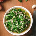 A white ceramic bowl of finished chimichurri sauce on a wooden board, showing the bright green herb mixture swimming in olive oil. Fresh parsley, shallots, and garlic cloves are visible in the background.