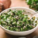 A close-up of a white bowl containing chimichurri sauce with a silver spoon, showcasing the texture of finely chopped herbs and red onions suspended in olive oil. In the background are steaks on a wooden board.