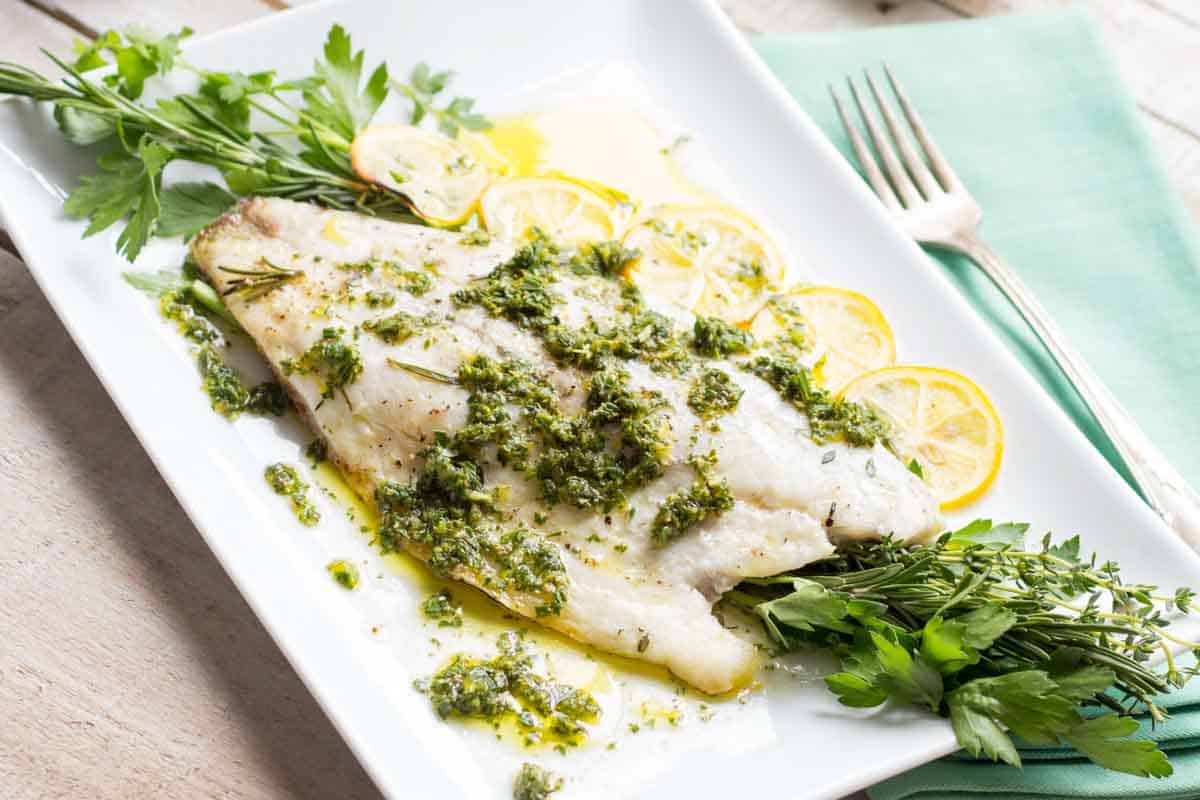 Baked white fish fillet on a rectangular white plate garnished with lemon slices, fresh herbs, and green herb sauce, with a fork on the side.