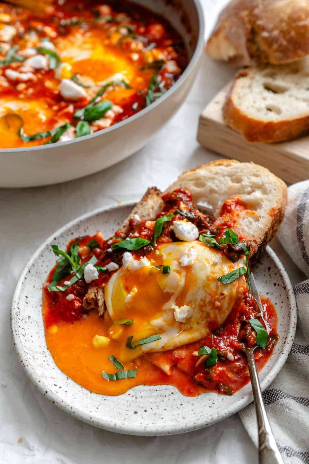 Close-up of a serving of eggs in purgatory on a speckled white plate with runny yolk being broken with a fork, garnished with basil and goat cheese, with skillet and bread visible in background.
